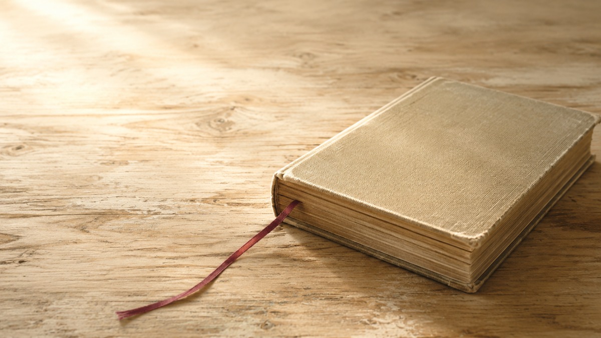 木の卓に置かれた布装の本から垂れる細い栞紐。紙の本に残る「どのあたりに書いてあったか」という場所の感覚のイメージ
