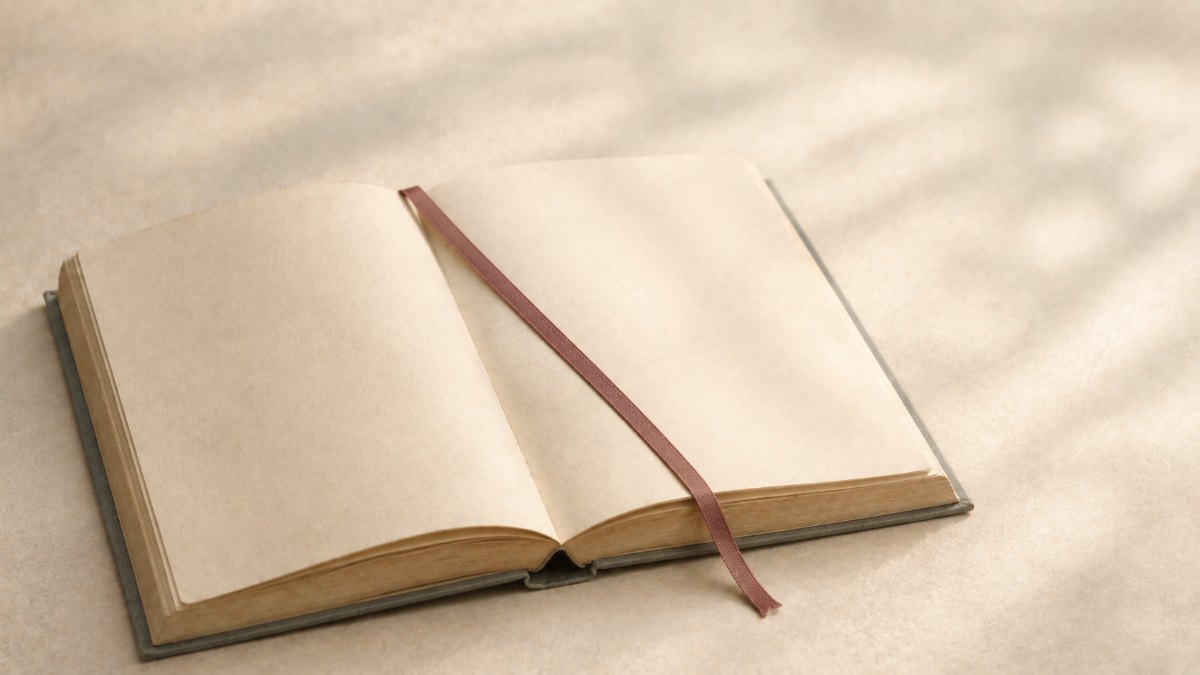 明るい日陰で開かれた無地の本のページに、細いリボンの栞が斜めに置かれている。どこから読むかを決めるイメージ