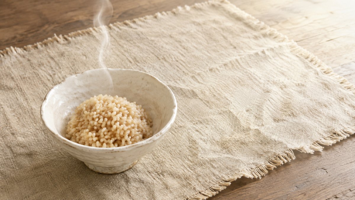 明るい昼の光の食卓に、麻のマットと粉引きの飯茶碗に盛られた玄米。茶色い炭水化物に置き換える健やかな一食のイメージ